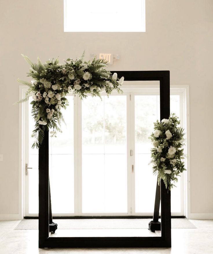 a black frame with flowers and greenery on the top is in front of a window