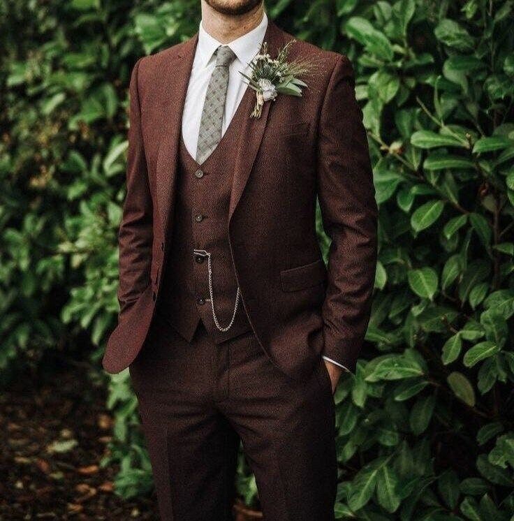 a man wearing a suit and tie standing in front of some bushes with his hands in his pockets