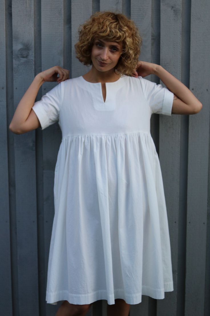 a woman wearing a white dress standing in front of a metal wall with her hands on her head