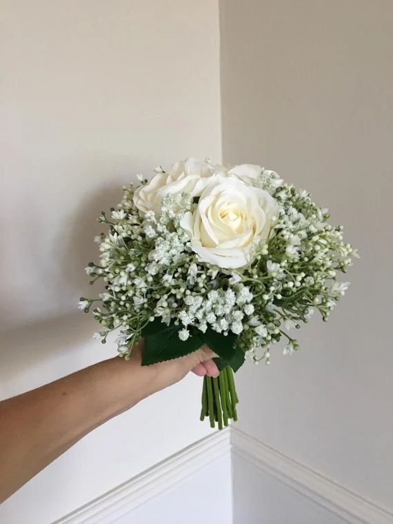 a hand holding a bouquet of white roses and baby's breath in front of a wall