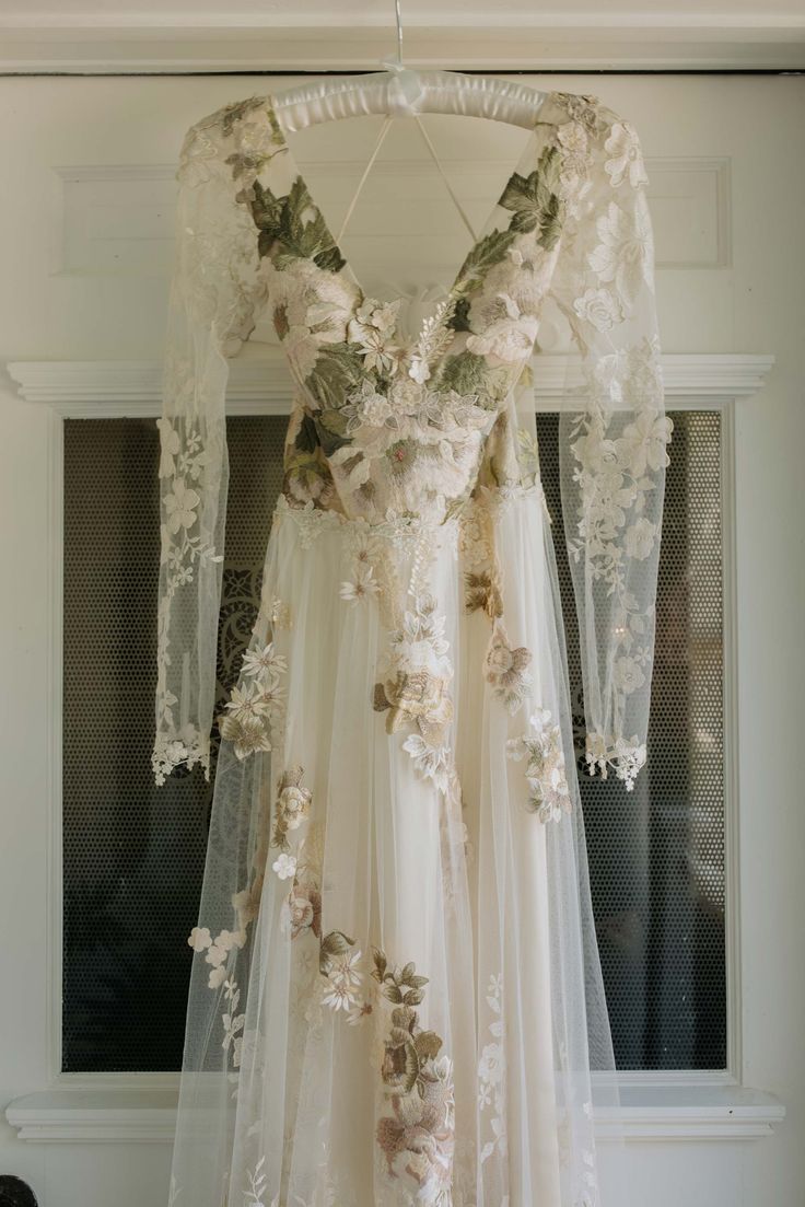 a white wedding dress with flowers on the back and long sleeves, hanging in front of a window