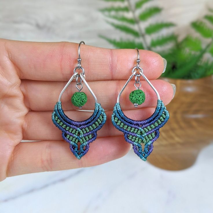 a hand holding a pair of green and blue beaded earrings in front of a potted plant