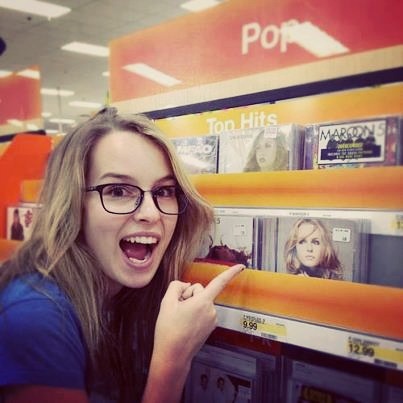 a woman in glasses pointing to the top hits sign on display at an electronics store