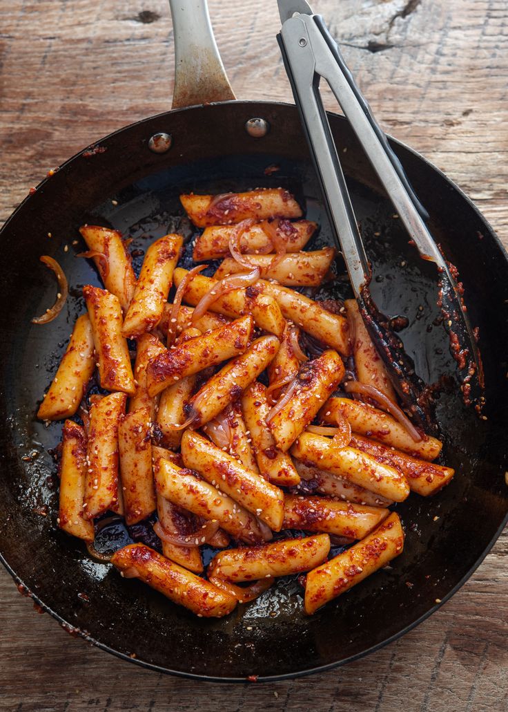 a frying pan filled with chicken wings and some tongs next to it on top of a wooden table