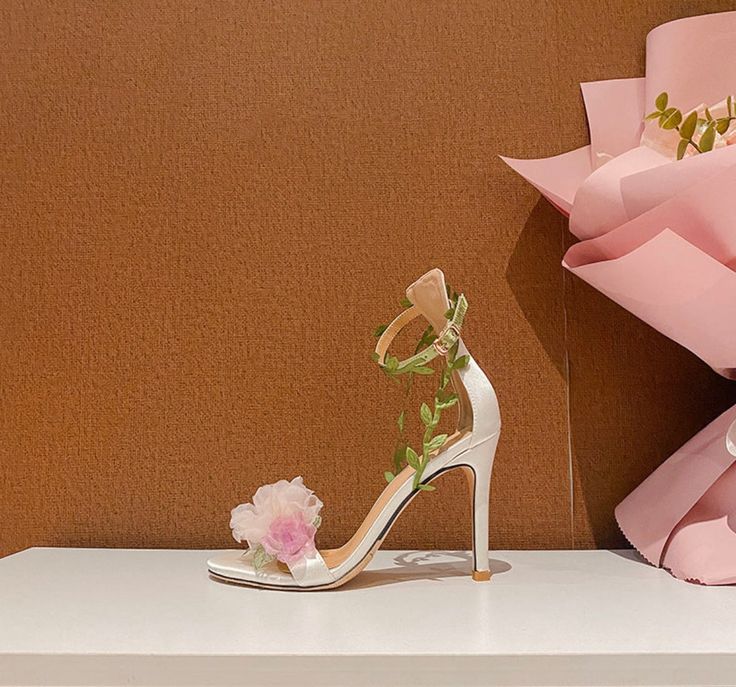 Step into a garden of elegance with the Claire Floral Heels. These stunning heels feature a delicate pink base adorned with intricate floral details, making them the perfect accessory for a romantic and feminine look. The ankle strap is beautifully decorated with green leaf accents, adding a whimsical touch that complements the soft, ethereal flowers on the toe. Crafted with a satin finish, the Claire Floral Heels offer a luxurious feel and a comfortable fit. The open-toe design showcases your p Pink Open Toe Wedding Shoes For Spring, Feminine Spring Wedding Shoes With Round Toe, Spring Wedding Guest High Heel Sandals, Spring Pink Wedding Shoes With Heel Strap, Spring High Heel Sandals For Wedding Guest, High Heel Sandals For Wedding Guests In Spring, Pink Spring Wedding Shoes, Chic Spring Wedding Shoes With Ankle Strap, Spring Wedding Heels With Wrapped Heel