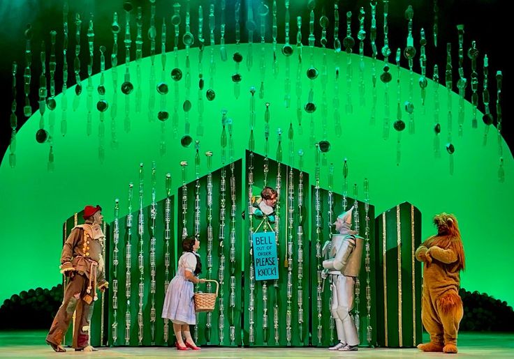 several people dressed in costumes stand on stage with green lights behind them and trees hanging from the ceiling