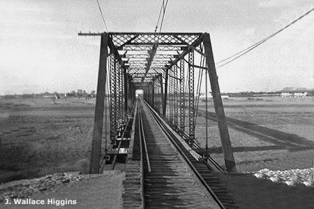 an old black and white photo of a train going over a bridge with power lines above it