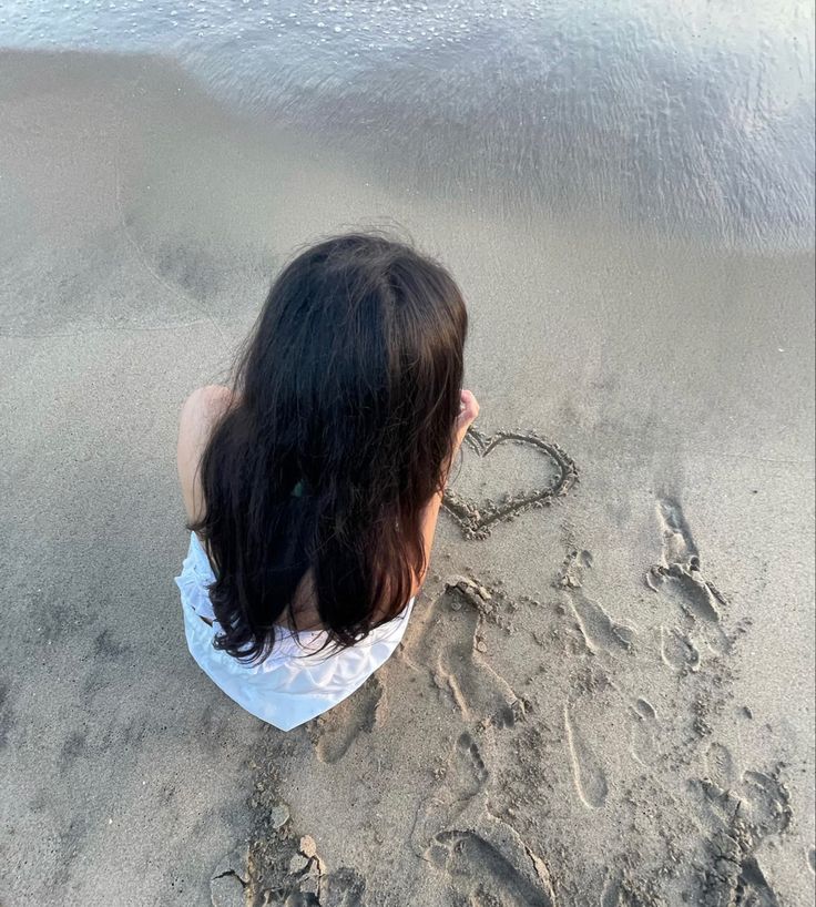 a woman sitting in the sand on top of a beach with a heart drawn in the sand