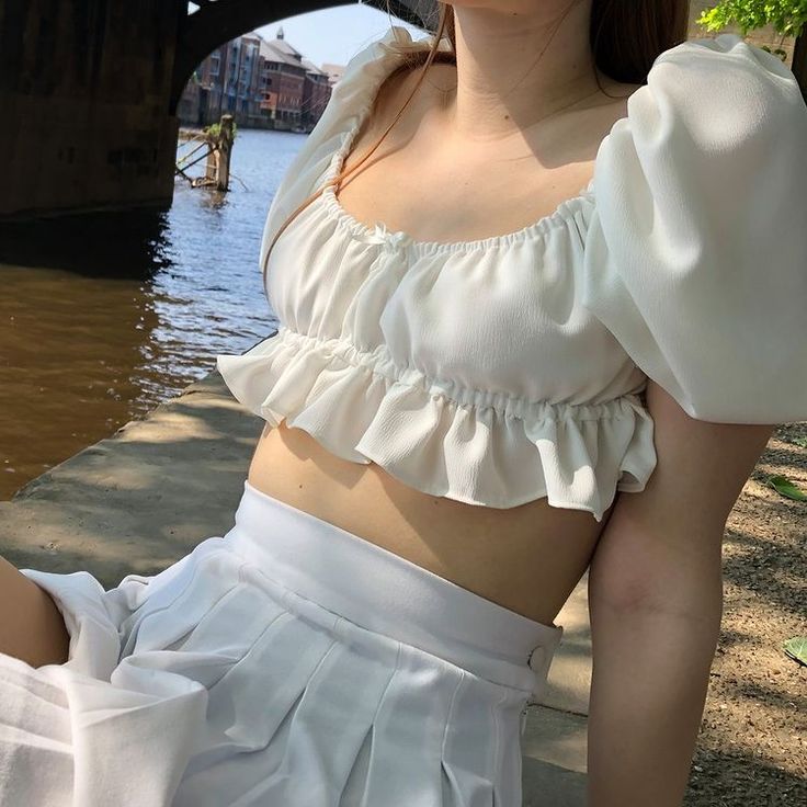 a woman wearing a white top and skirt posing in front of a river under a bridge