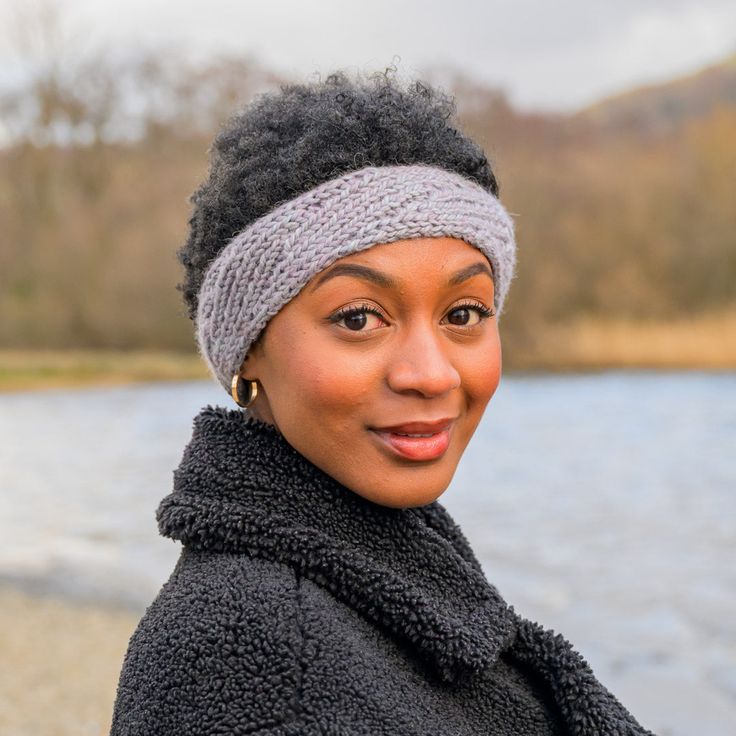 a woman wearing a knitted headband and smiling at the camera with a body of water in the background