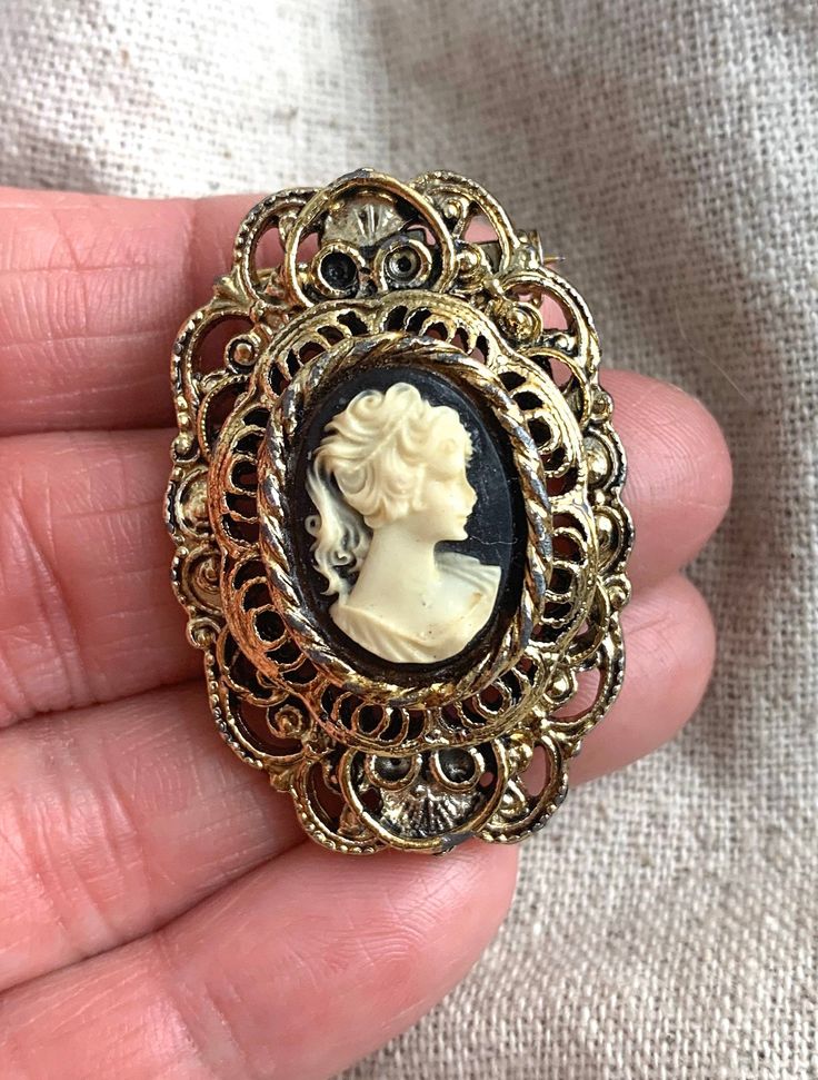 "You will receive this vintage cameo brooch from Gerry's. It measures 1 3/4\" x 1 1/8\" and is cast in an antiqued gold tone base metal. The cameo backing is black and the silhouette is a cream color. This brooch is in very good vintage condition with minimal signs of wear and a properly functioning safety catch. For finished vintage assemblage and vintage inspired necklaces, bracelets, and earrings, please visit us at our other Etsy store, William Dalton Design: www.williamdaltondesign.etsy.com Vintage Medallion Brooch For Wedding, Vintage Medallion Brooches For Wedding, Ornate Medallion Brooches For Collectibles, Ornate Medallion Brooches For Collectors, Antique Cameo Medallion Brooch, Antique Cameo Medallion Brooches, Antique Gold Brooches For Vintage Collection, Antique Gold Brooches From Vintage Collection, Gold Antique Brooches For Vintage Collection