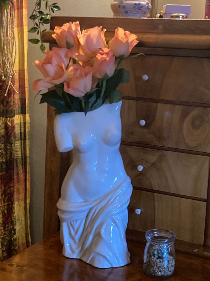 a white vase with pink roses in it on a table next to a dresser and mirror