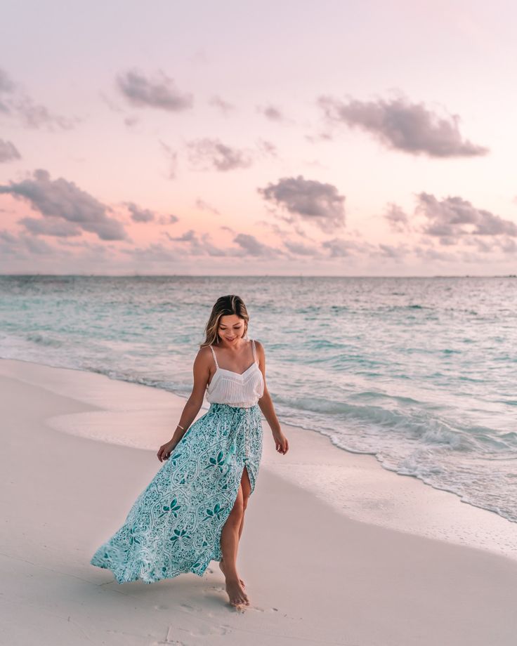 a woman standing on top of a sandy beach next to the ocean at sunset with her dress blowing in the wind