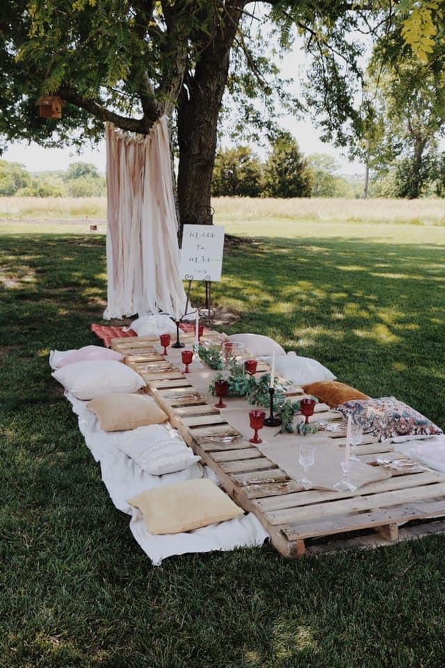 an outdoor table set up in the grass with candles and napkins on it, next to a tree