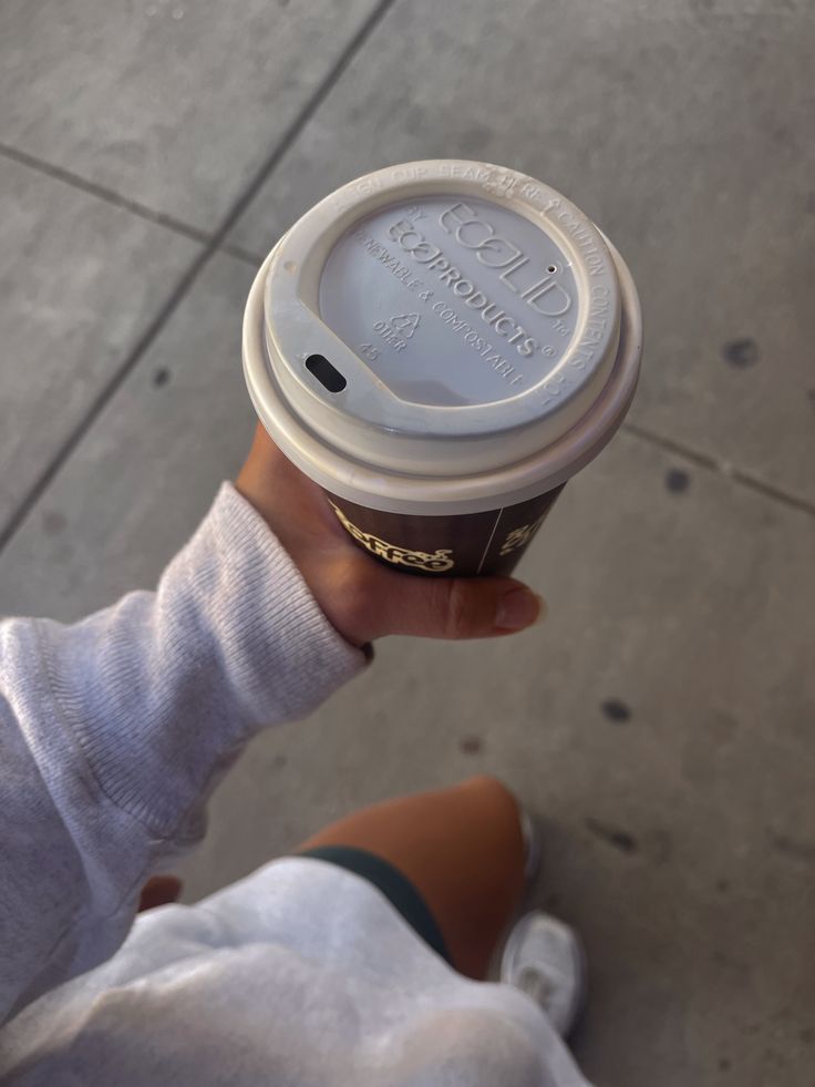 a person holding up a cup of coffee on the sidewalk with their thumb in it