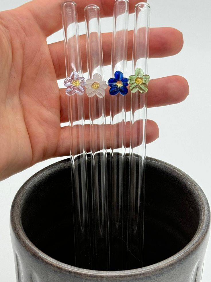 a hand is holding five clear plastic tubes with flowers on them in front of a black cup