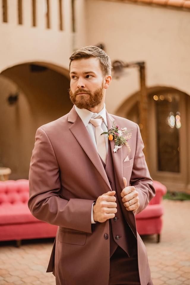 a man in a suit standing next to a pink couch and red sofa with his hand on his tie