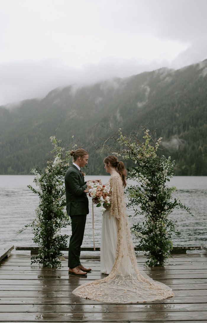 a bride and groom standing on a dock next to the water with mountains in the background