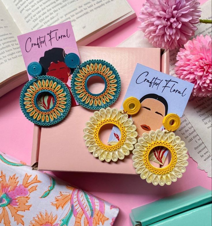 several colorful earrings are laying on top of a book and some pink flowers in the background