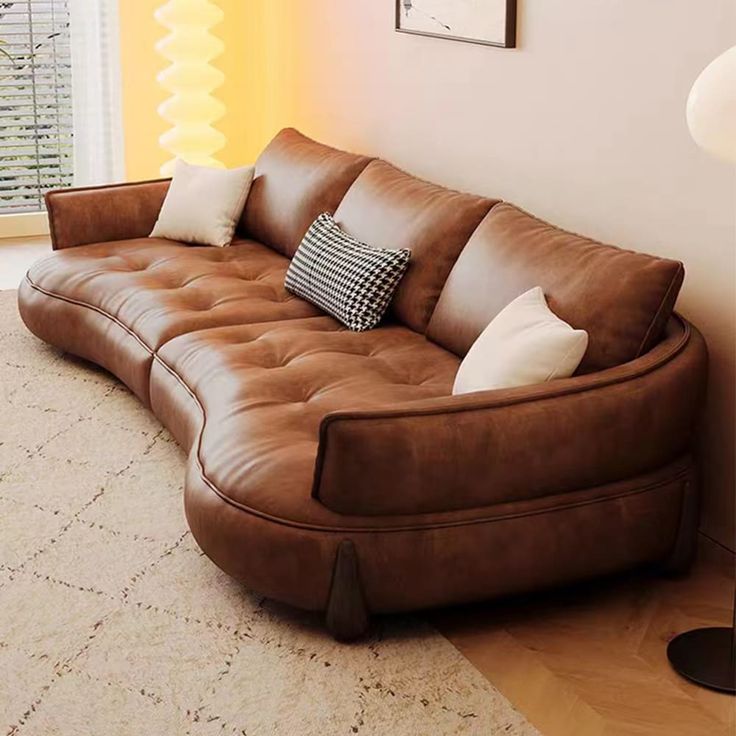 a brown leather couch sitting in front of a window next to a lamp and pictures on the wall