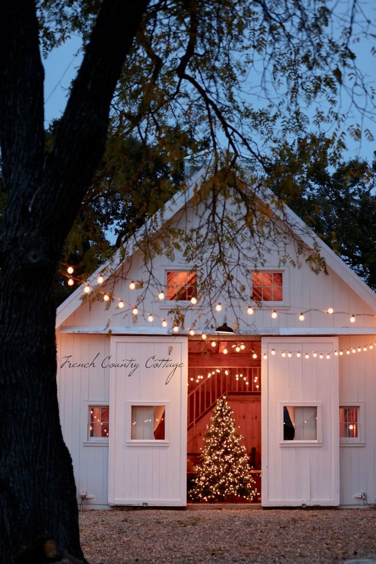 a christmas tree is lit up in front of a white barn with lights on it