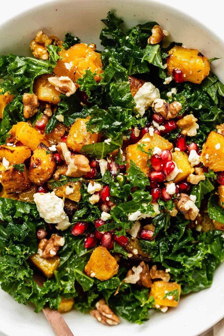 a white bowl filled with kale salad topped with pomegranate, nuts and feta cheese