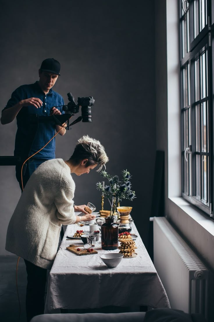 a man and woman preparing food on a table in front of a window with a camera