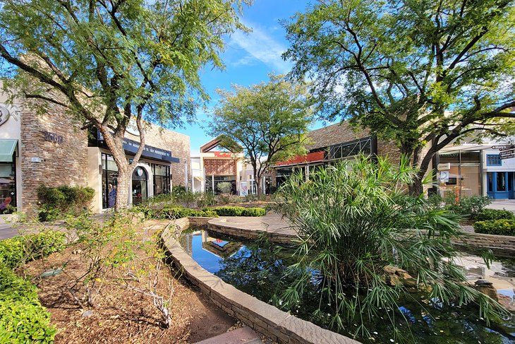 a small pond in the middle of a street with buildings and trees around it, surrounded by greenery