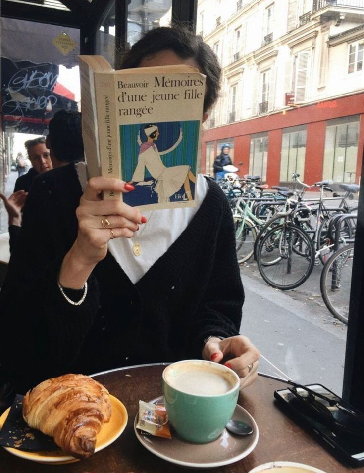 a woman sitting at a table reading a book and eating croissants in front of her