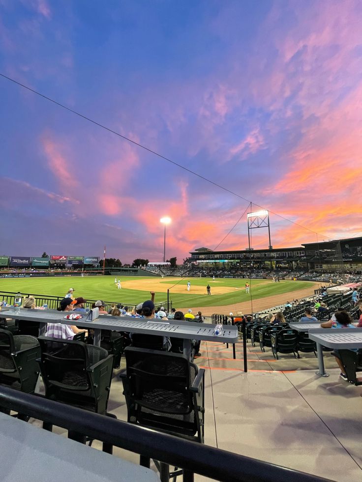 a baseball field with people sitting at tables and watching the sun go down in the distance