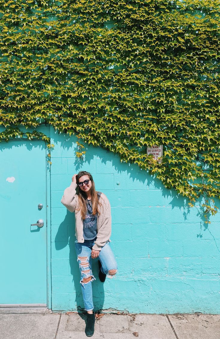 a woman leaning against a blue wall with ivy growing on the side and wearing ripped jeans