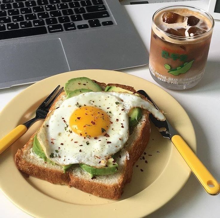 an egg and avocado toast on a plate next to a drink with a laptop in the background