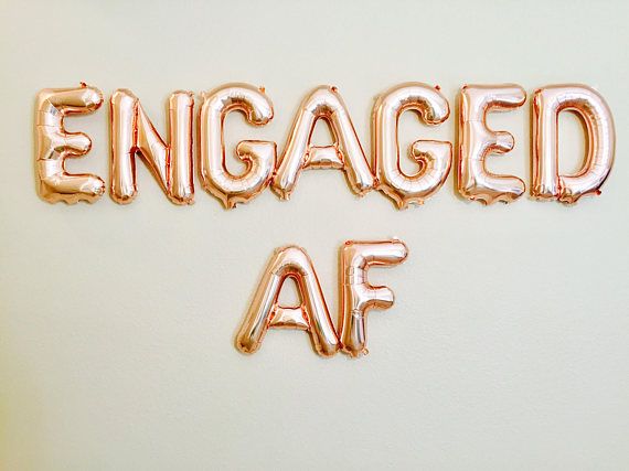 the word engaged af spelled out in gold foil letters on a white wall with balloons