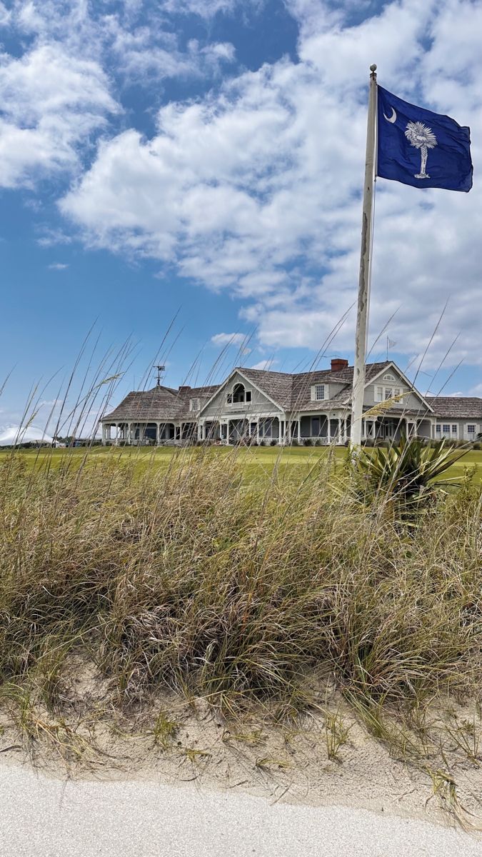 a flag flying in front of a large white house on the beach with tall grass