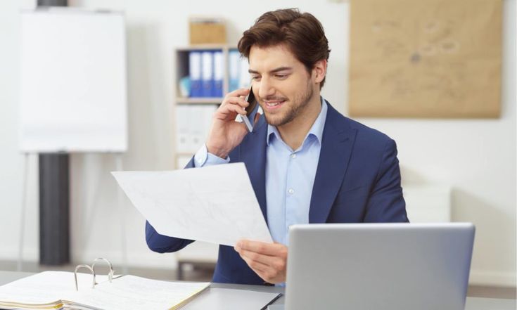 a man sitting at a desk talking on the phone and holding a piece of paper