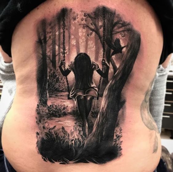 a man with a tattoo on his back is sitting in the woods and holding onto a swing