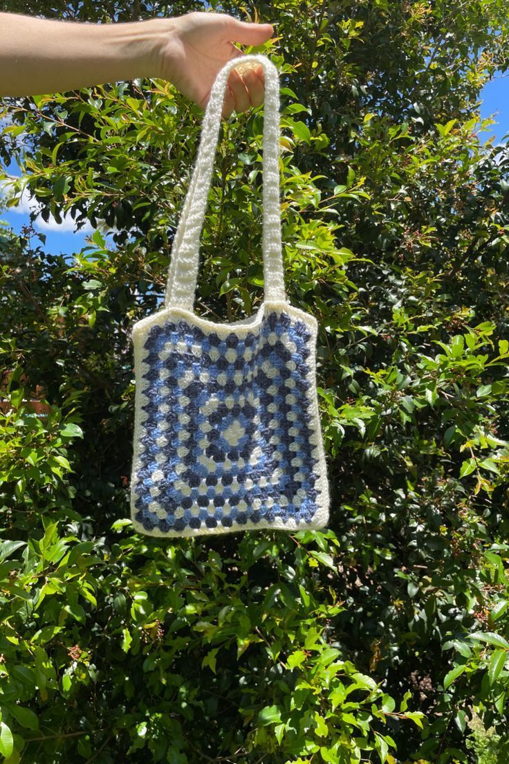 a hand is holding a crocheted bag in front of some trees and bushes