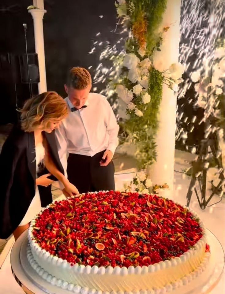 a man and woman standing in front of a large cake with red flowers on it