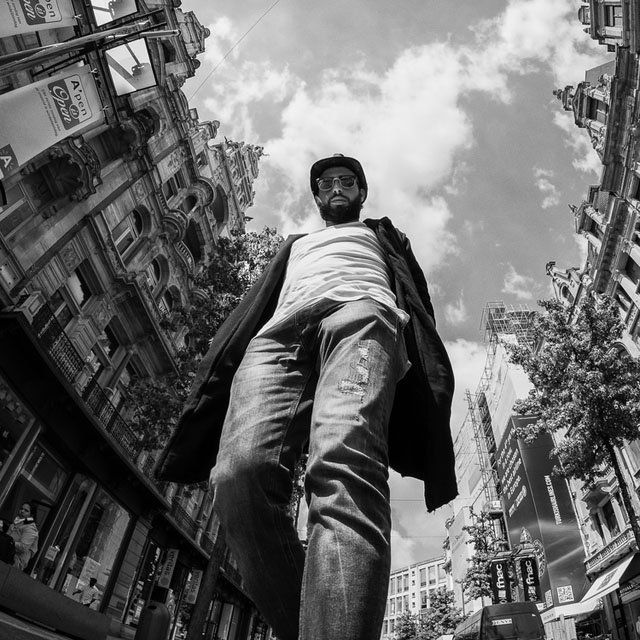 a man is walking down the street in front of some tall buildings and trees with his head tilted to the ground