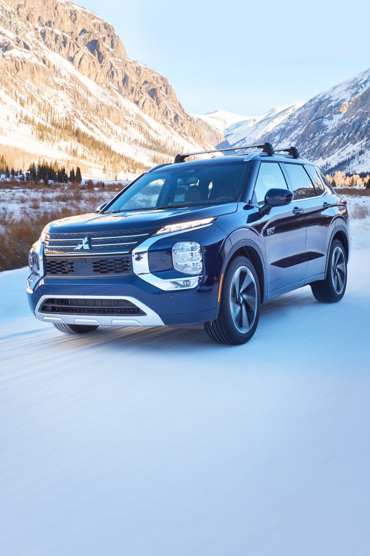 a blue suv driving down a snow covered road in the middle of winter with mountains in the background