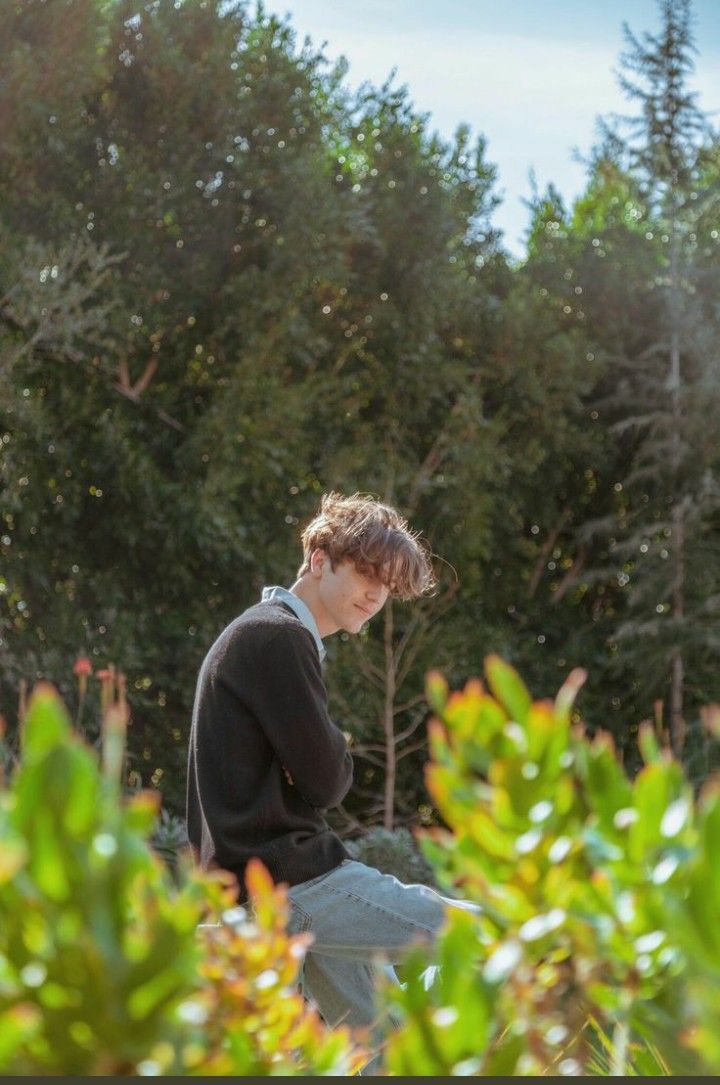 a young man sitting on top of a wooden bench in front of some bushes and trees