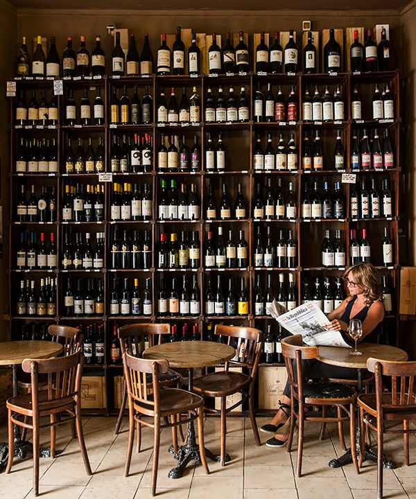 a woman sitting at a table reading a newspaper in front of a wall full of wine bottles