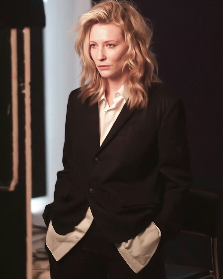 a woman in a black suit and white shirt standing next to a chair with her hands on her hips