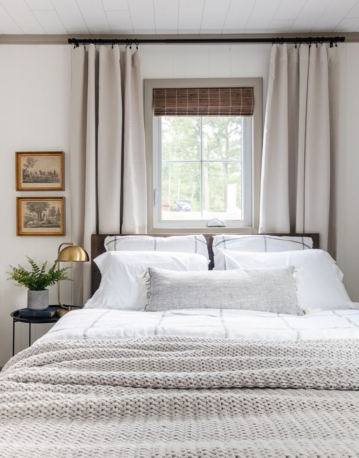 a bed with white sheets and pillows in a bedroom next to a window filled with curtains