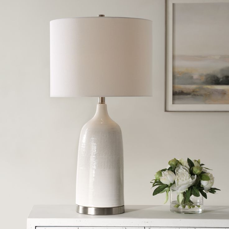 a white table lamp sitting on top of a dresser next to a vase with flowers