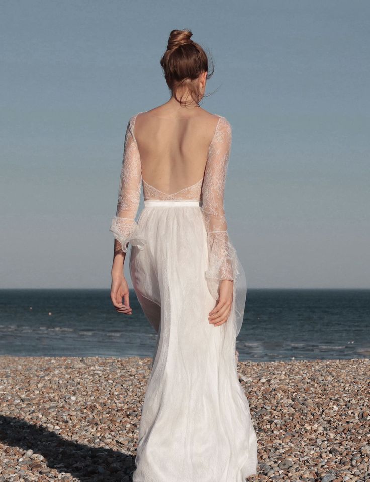 the back of a woman in a white wedding dress standing on rocks near the ocean