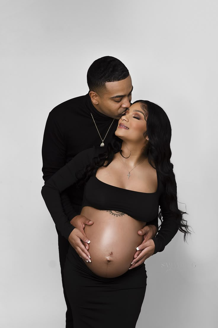 a pregnant couple cuddles and kisses while posing for a photo in front of a white background