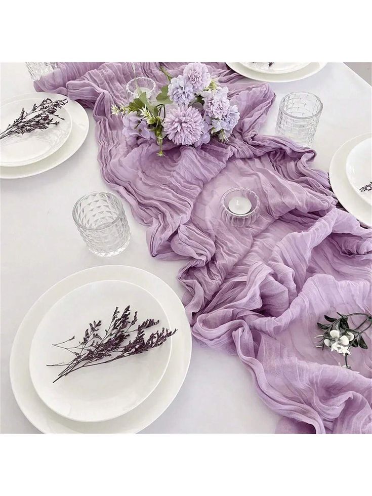 the table is set with white plates and purple cloth on it, along with lavender flowers