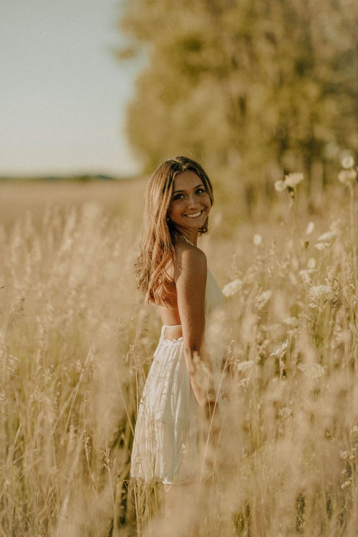 a woman standing in the middle of a field with tall grass and trees behind her