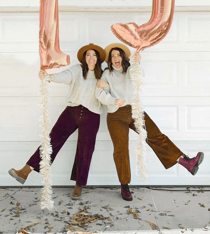 two women wearing hats and holding balloons in front of a garage door with their arms around each other